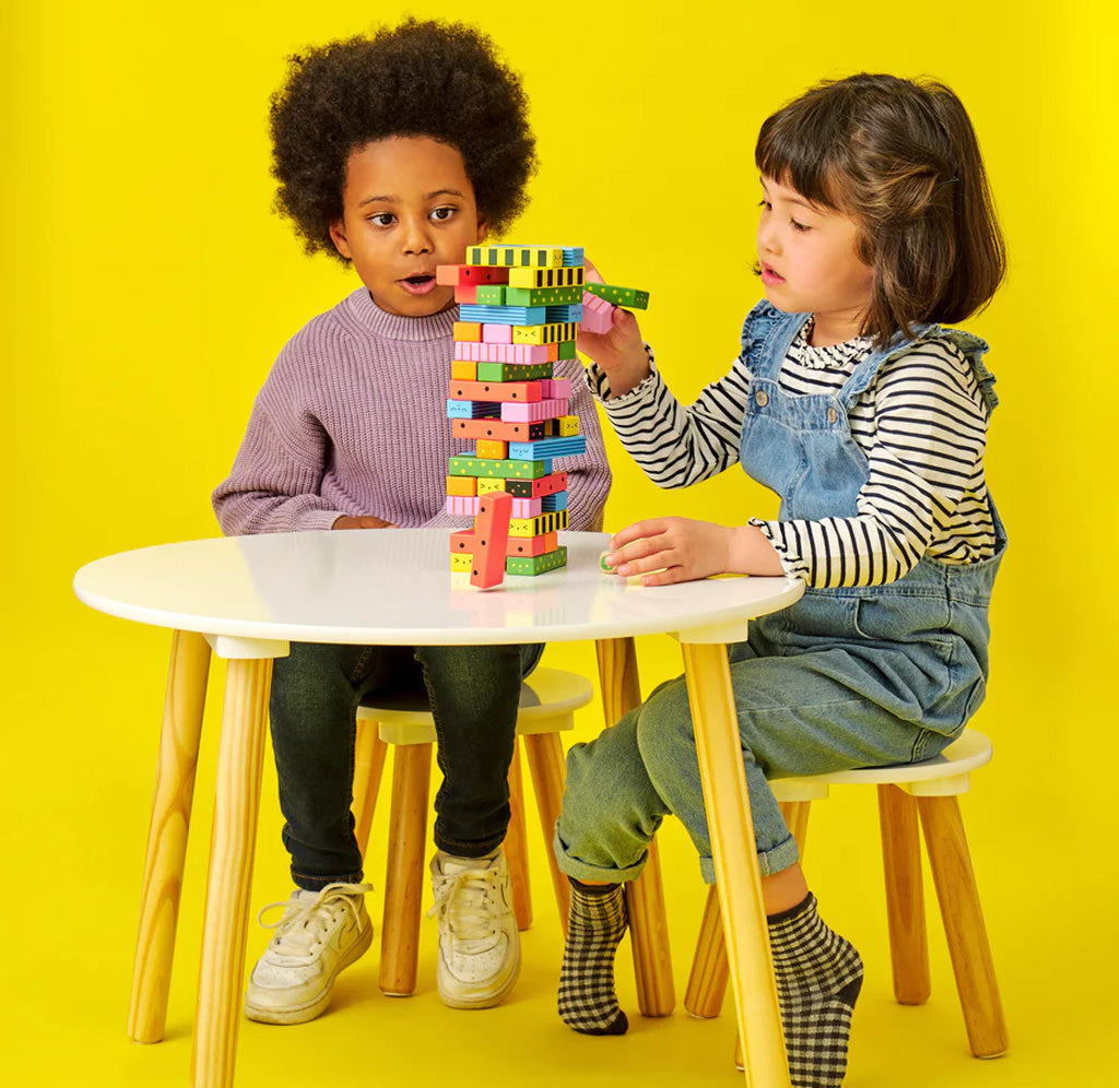 Two children sitting at a table playing with multi-colored wooden blocks stacked up in a column. One child is pulling a block from the tower while the other kid watches. 