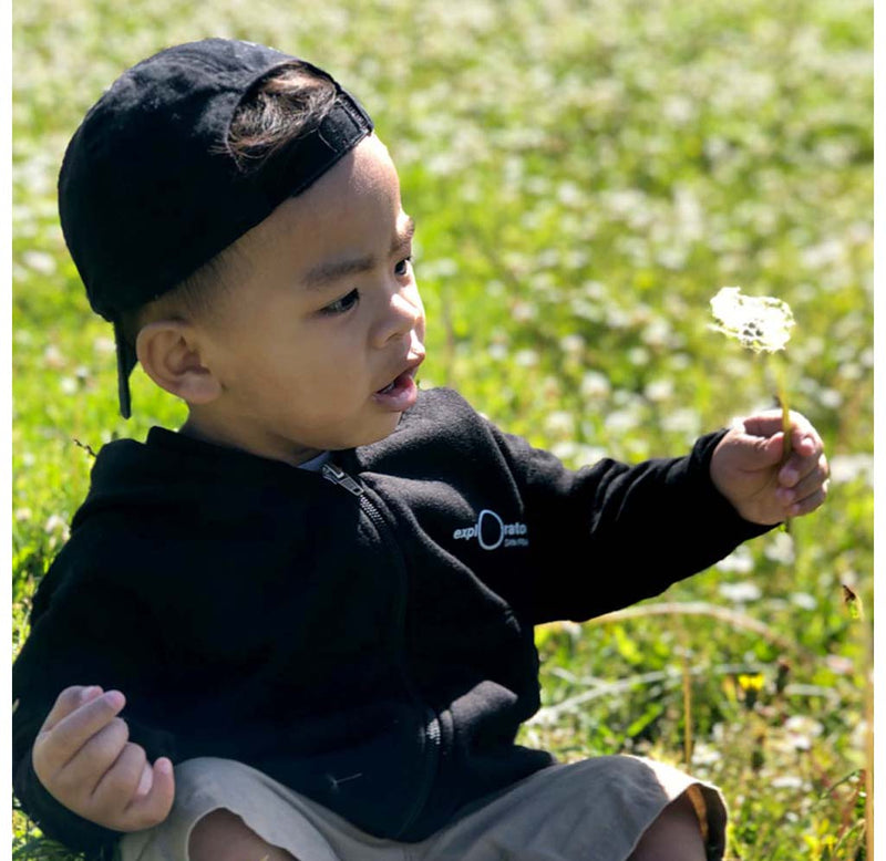 Young child sitting on the grass wearing a black hoodie with Exploratorium on the front in white.