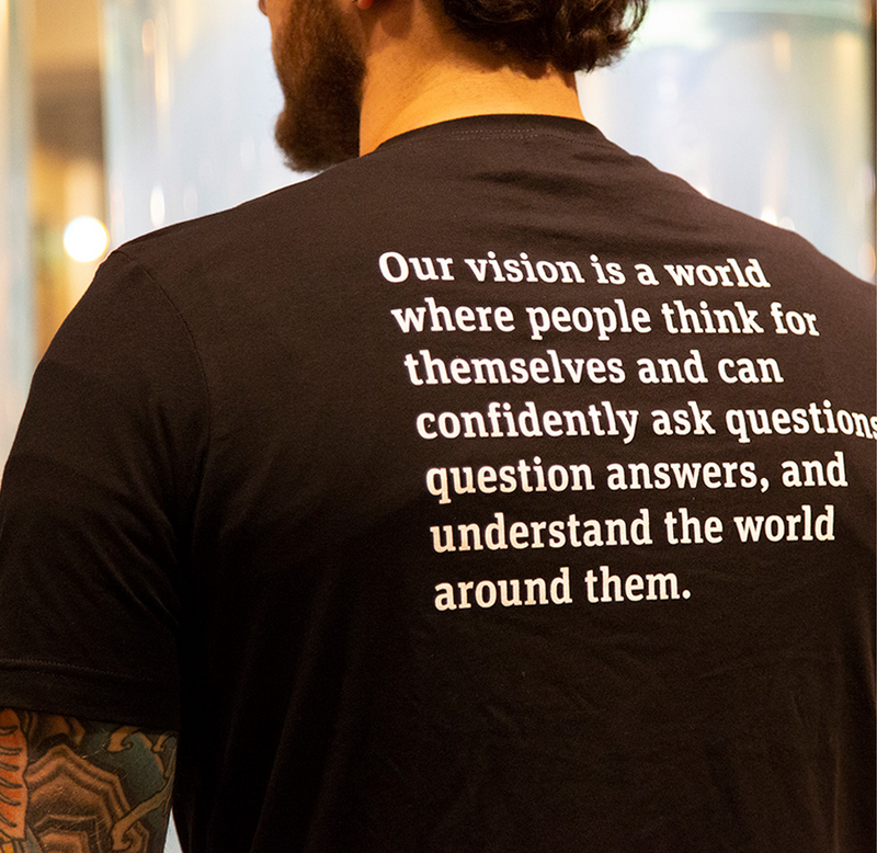 A male is standing with his back facing forward; he is wearing a black t-shirt with the Exploratorium's mission statement on the back in white. It reads, "Our vision is a world where people think for themselves and can confidently ask questions, question answers, and understand the world around them."