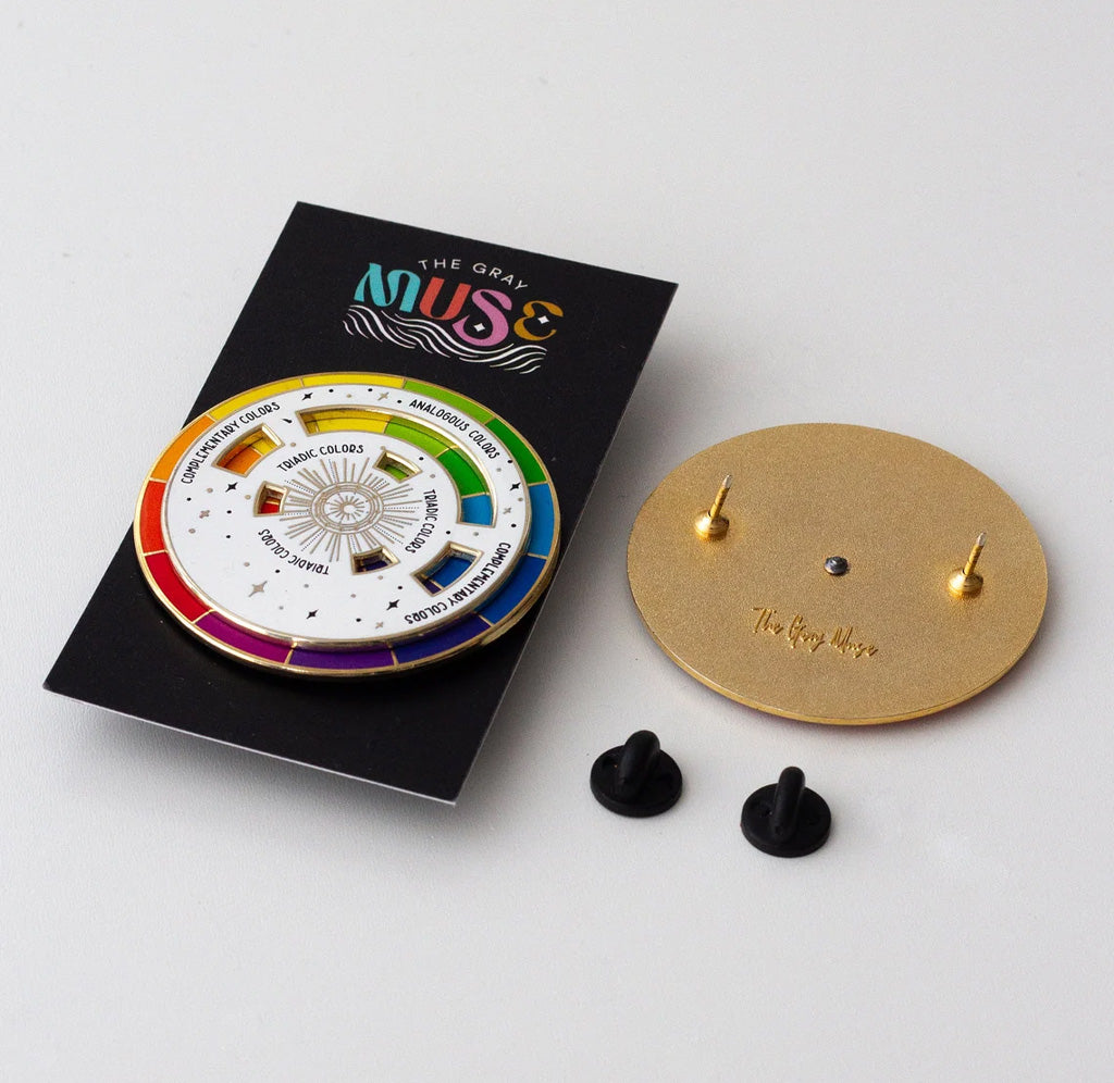 Enamel pin displayed on a black cardboard backing. Next to the packaged pin, there is another pin flipped so the back is showing, with a gold plated back and two pin posts attached. Two rubber backings sit below the overturned pin. 