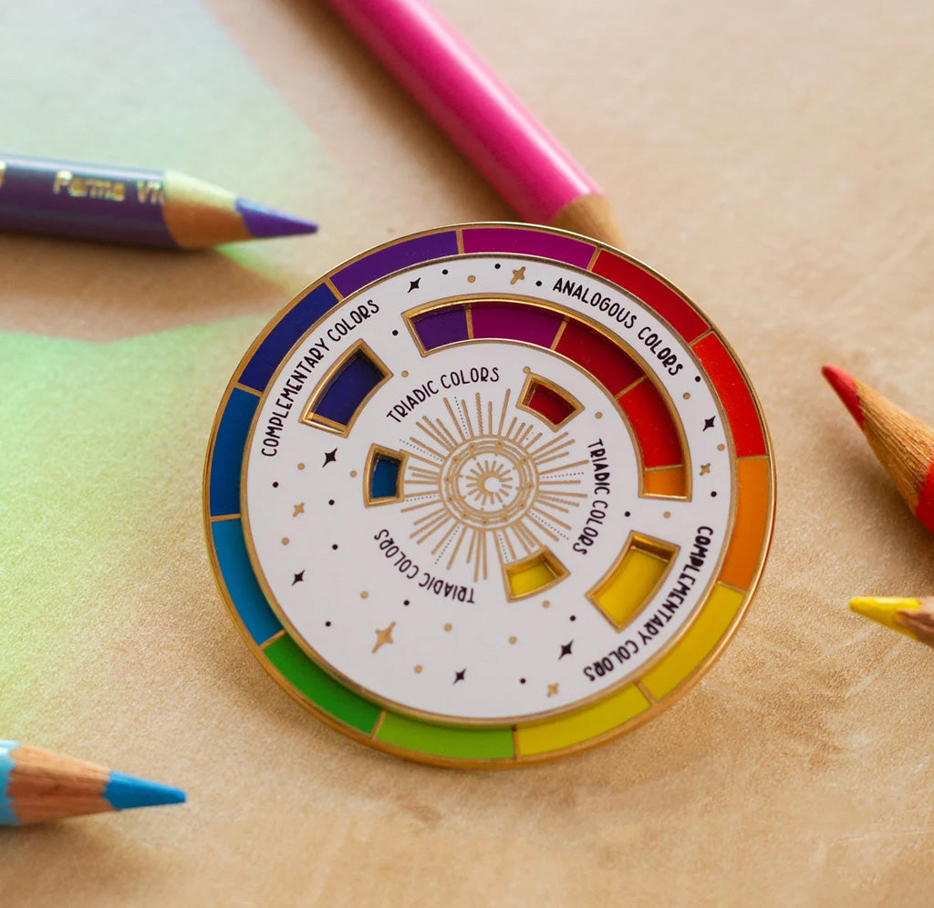Round enamel pin outlined in gold. Two circles make up the pin, with the outer circle displaying 12 colors in a wheel. The inner circle has cutouts that show a sub section of the color wheel. Pin is sitting on a wooden table with colored pencils in the background. 