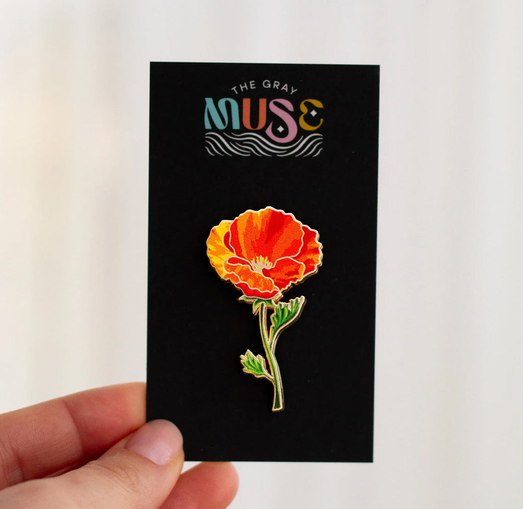 An orange and red colored poppy flower enamel pin on a black cardboard backing. 