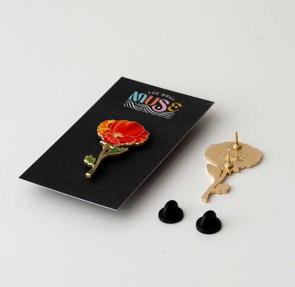 Angled view of the poppy enamel pin, with the cardboard-backed pin to the left and the pin facing down on the table to the right. The back of the pin is gold and has two sharp points secured near the top of the pin.  Two black rubber pin fasteners are on the table below the turned over pin.