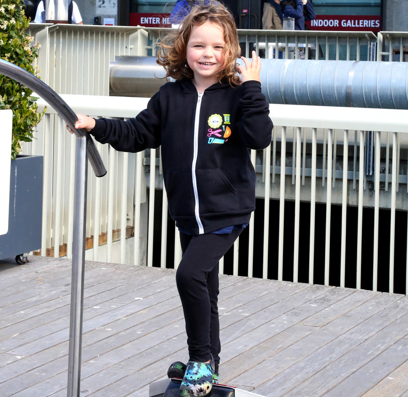 A young child is standing on a pier standing on an exhibit. She is wearing the black Art of Tinkering hoodie with brightly colored neon tools in pink, orange, teal, green and yellow.