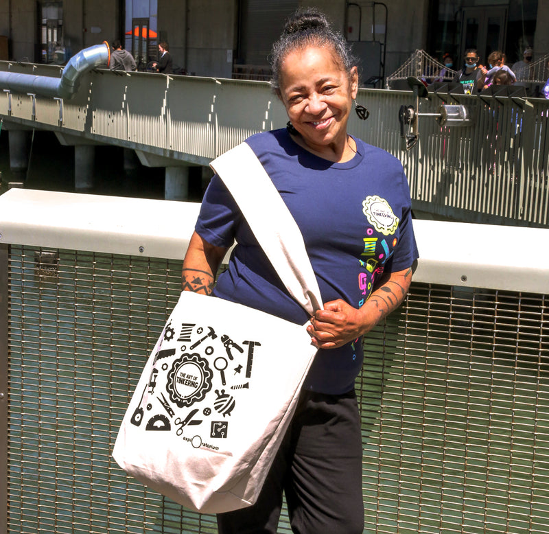 A woman is standing on the bridge, wearing the navy blue with brightly colored neon tools and Art of Tinkering logo t-shirt and holding the white canvas silked screened in black with tools Art of Tinkering tote bag.