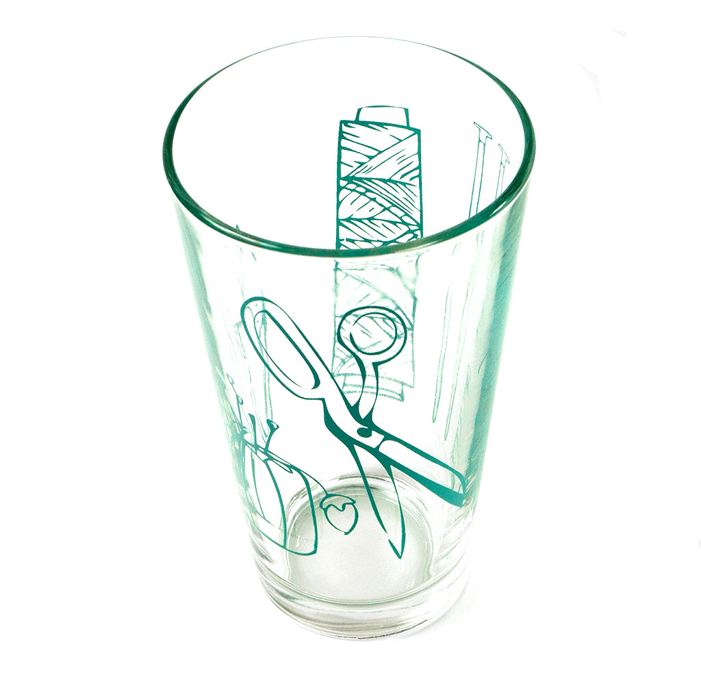  A 16oz pint glass with scissors, snippers, pincushion, knitting needles, crochet hook, thread spool, and cone are screen printed all around in a turquoise silhouette.