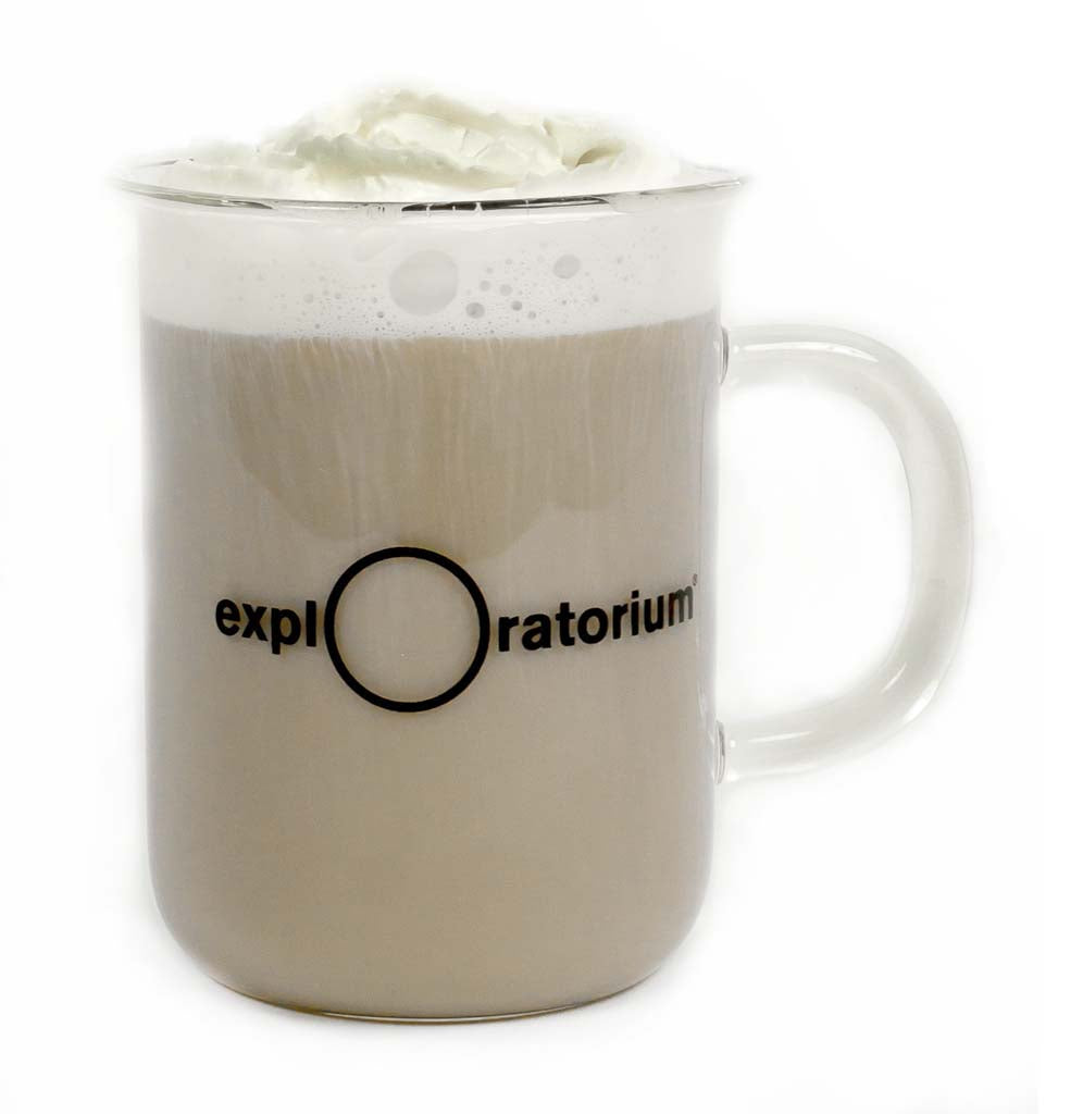 A clear glass beaker mug filled with a latte with the Exploratorium logo in black across the front. 
