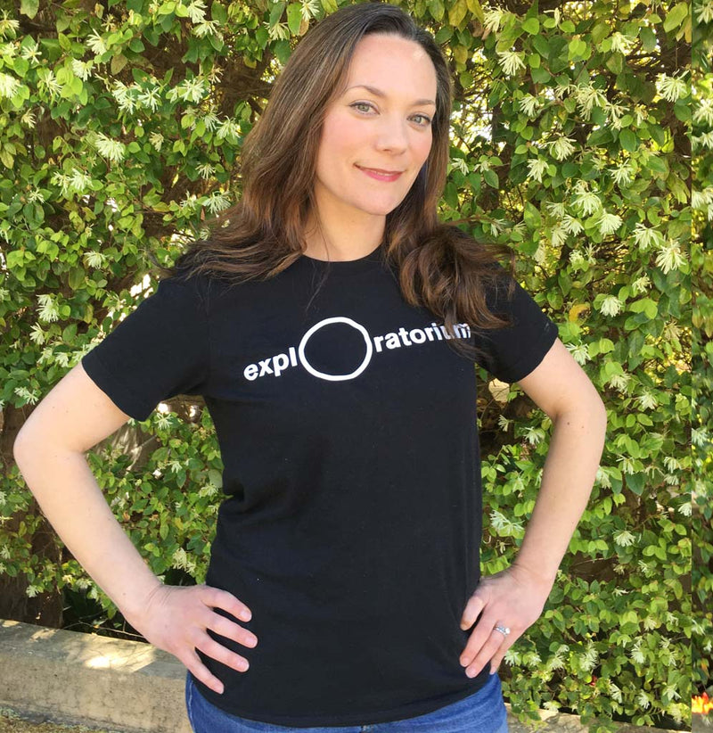A woman is standing facing forward; she is wearing a black t-shirt with the Exploratorium in white across the front of the shirt.