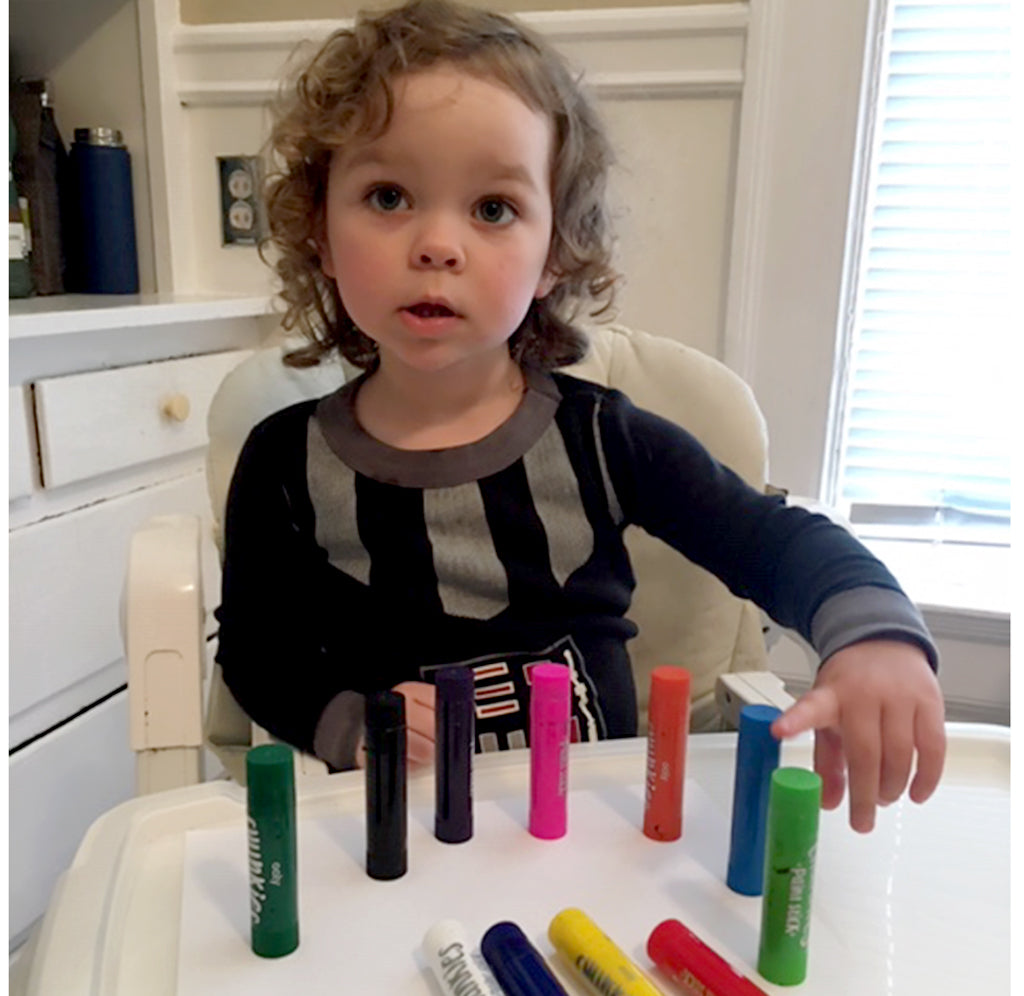 A small child sits at a table with different colored paint sticks standing and lying on the table.