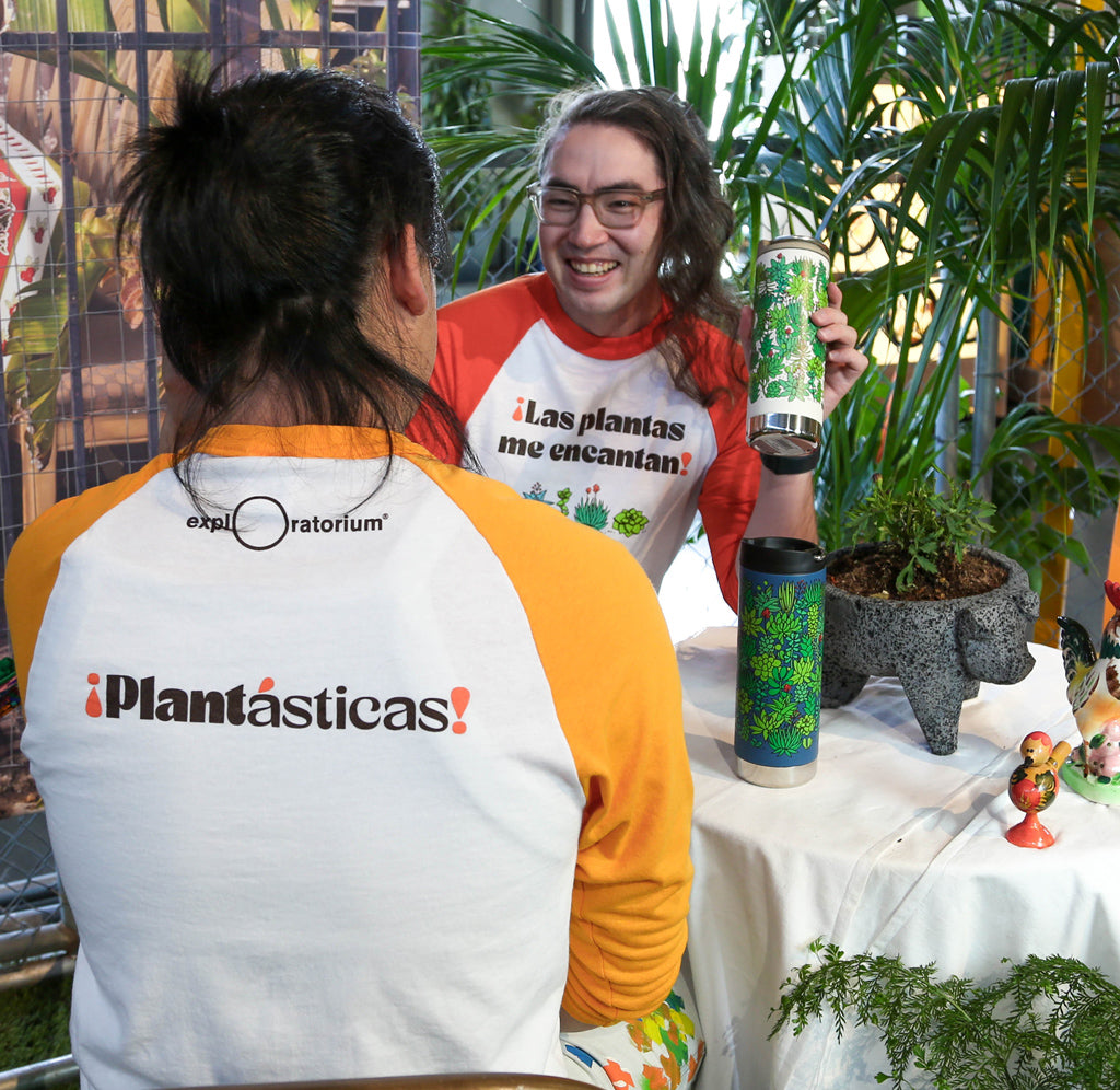 Two people seated wearing the Exploratorium baseball tee shirt  that has ¡Las plantas me encantan! printed on the front and Exploratorium and ¡Plantasticas! printed on the back. 