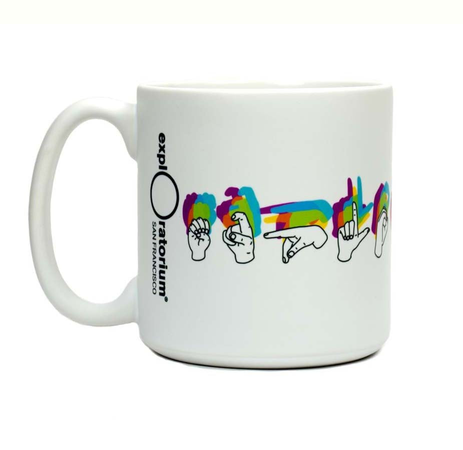 A white ceramic mug with Exploratorium San Francisco printed vertically next to the handle. Exploratorium follows it spelled out horizontally around the mug in sign language. The signed letters cast a multi-colored shadow behind them. 