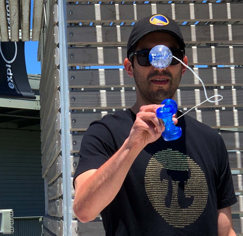A gentleman is standing while playing with a blue light-up Kendama. The Pi shirt is black with gold ink in 1,878 digits of Pi surrounding a blank Pi symbol. 