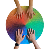A large circular jigsaw puzzle featuring a rainbow spectrum of colors arranged in radial segments, photographed from above on a white background with four hands resting on top.