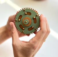 Hands hold a round green mechanical rotor with copper coils, brass contacts, and toothed edges, creating a detailed, technical mood.