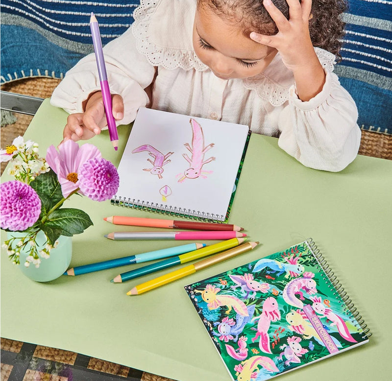 A young child is sitting at a table drawing pink axolotls with another sketchpad and different colored pencil sit on a green table top.