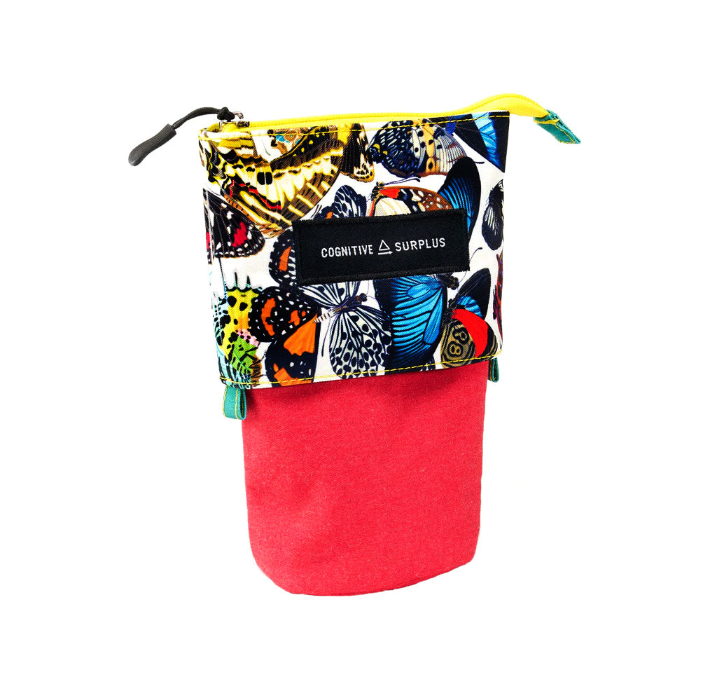 A small zippered pouch with a colorful butterfly print on the upper half and a solid red felt lower half, featuring a black label that reads 'COGNITIVE SURPLUS'.