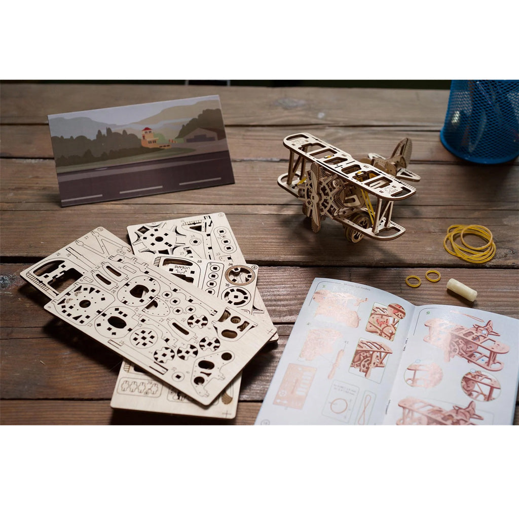 Environmental shot of the biplane model kit showing the wooden sheets containing the pieces to build the model, an instruction book, rubber bands, and wax. 