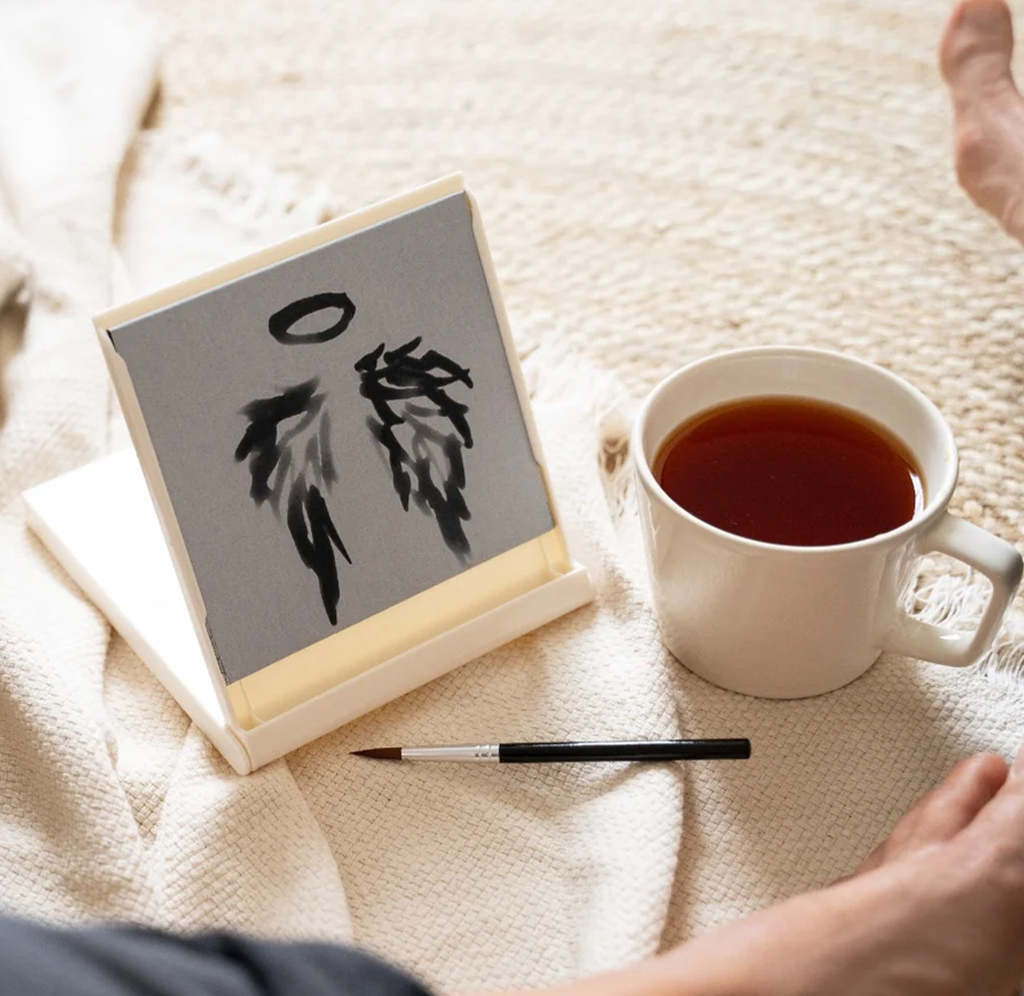 A small square canvas or print with a black feather-and-halo artwork resting on a cream blanket beside a white mug of tea and a paintbrush. A person's hands and feet are visible around the scene, indicating a cozy home environment.