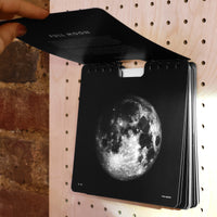 A spiral-bound black calendar with a large white moon print on the cover, being opened by a hand against a pegboard background.
