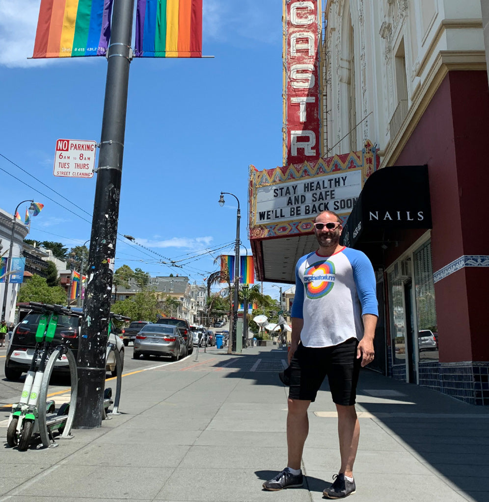 A male is standing on Market Street in San Francisco's Castro district. He is wearing a white long sleeve jersey with blue sleeves and a red collar with a circular rainbow design. Exploratorium is underneath in blue.