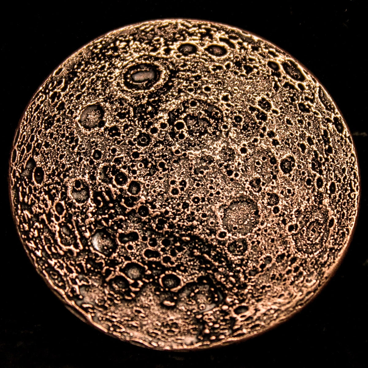 A circular copper-colored metal coin featuring a textured, cratered moon or planetary surface design, shown against a solid black background.