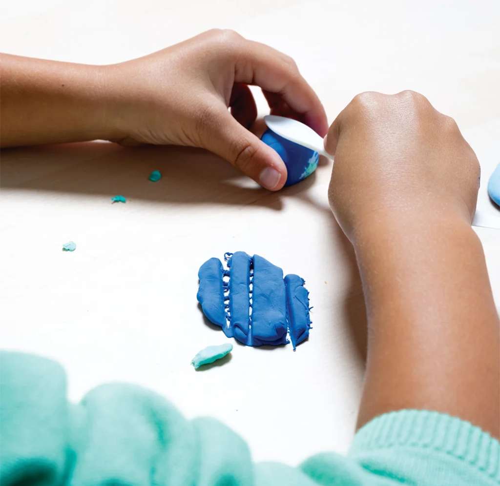 A child’s hands shape blue and turquoise modeling clay on a white table, with small clay pieces scattered nearby; a calm, creative activity.