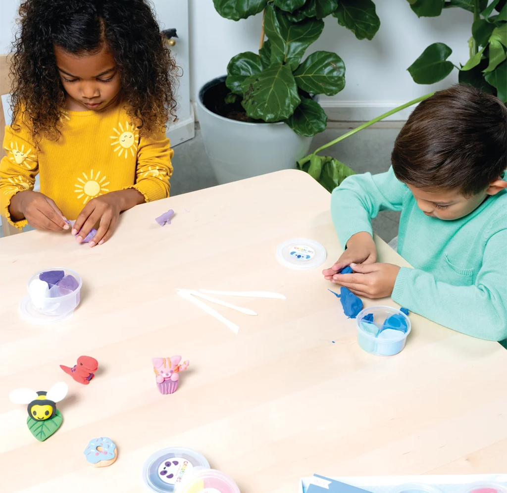 Two children in yellow and turquoise shirts shape colorful clay at a table, with small creations and tubs of clay nearby; the scene feels calm and creative.