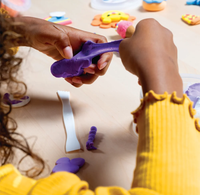 A child in a yellow sweater stretches purple modeling clay by hand, with plastic tools and colorful clay pieces scattered nearby in a playful scene.