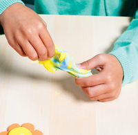 A child in a turquoise sweater stretches bright yellow-and-blue slime between both hands over a pale table, conveying playful, hands-on creativity.
