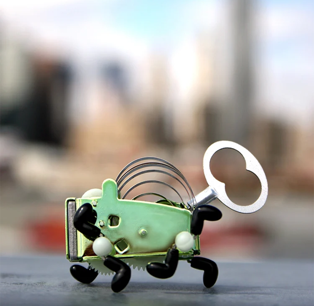 Whimsical green wind-up toy bug with black legs, white accents, coiled metal springs, and a large silver key, against a blurred cityscape.