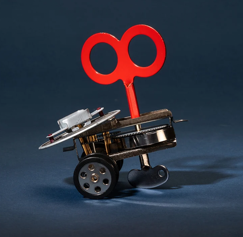 Whimsical wind-up toy vehicle with a bright red key, metal gears, wheels, and a small seat, staged against a dark blue background.