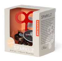 Open cardboard box displays a red wind-up mechanical creature toy with exposed gears; “SPARKLZ” packaging conveys a playful, whimsical tone.