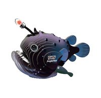 A DIY  cardboard anglerfish 3D kit put together against a  plain white background. The fish body is dark with iridescent purple and blue tones, white line graphics, a black eye, a small fishing line reel on the top, and a green lure attached near the tail.