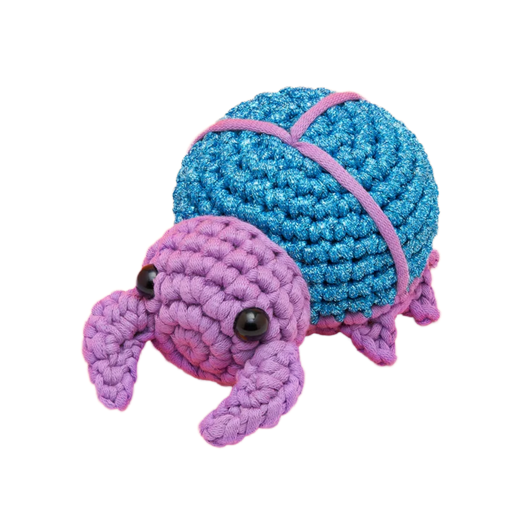 A handmade crocheted beetle with a bright blue, rounded shell crossed by pink straps, a purple head and flippers, and glossy black eyes, set against white. Its soft, playful look feels cheerful.