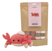 A crochet kit with coral, cream, and white yarn in a kraft-paper pouch, beside a finished smiling crab plush with raised claws; playful, cozy tone.