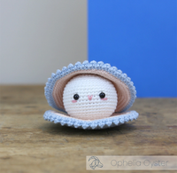 A shy crocheted oyster doll with a cream face, pink cheeks, and a pale blue ruffled shell sits on a wooden table against a blue-and-gray wall.