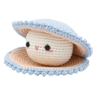 A cute crocheted cream-colored face with glossy black eyes and rosy cheeks peeks from a pale blue, peach-lined oyster shell, conveying a sweet, playful mood.