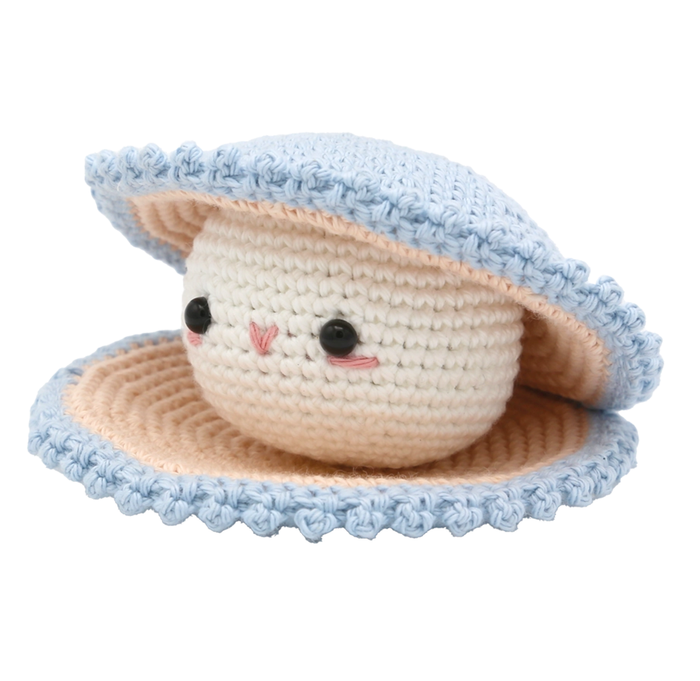 A cute crocheted cream-colored face with glossy black eyes and rosy cheeks peeks from a pale blue, peach-lined oyster shell, conveying a sweet, playful mood.