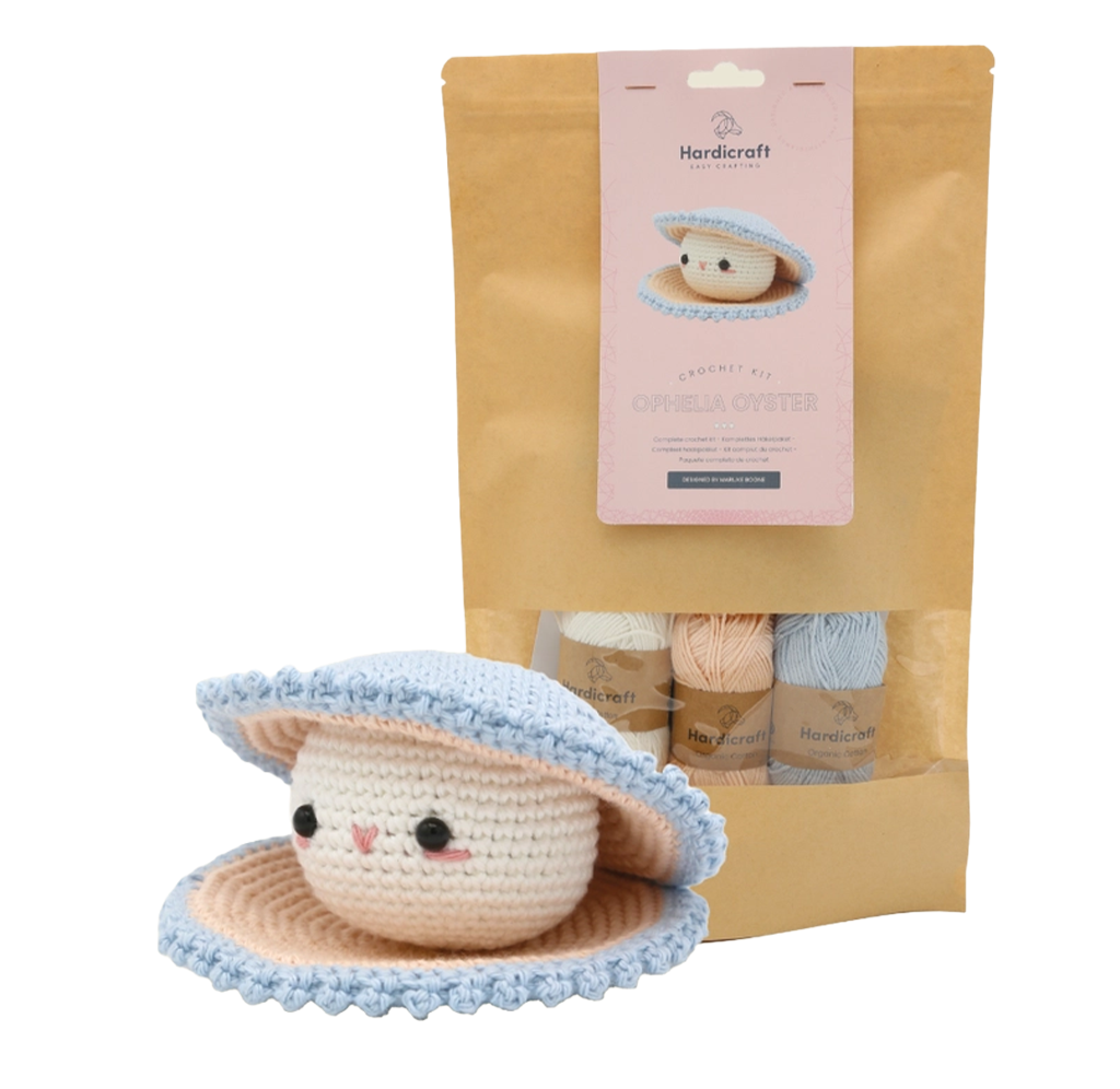 A handmade crochet oyster with a smiling face and blue-and-cream saucer sits beside its craft kit, packaged in a kraft bag with yarn skeins; cozy and cheerful.