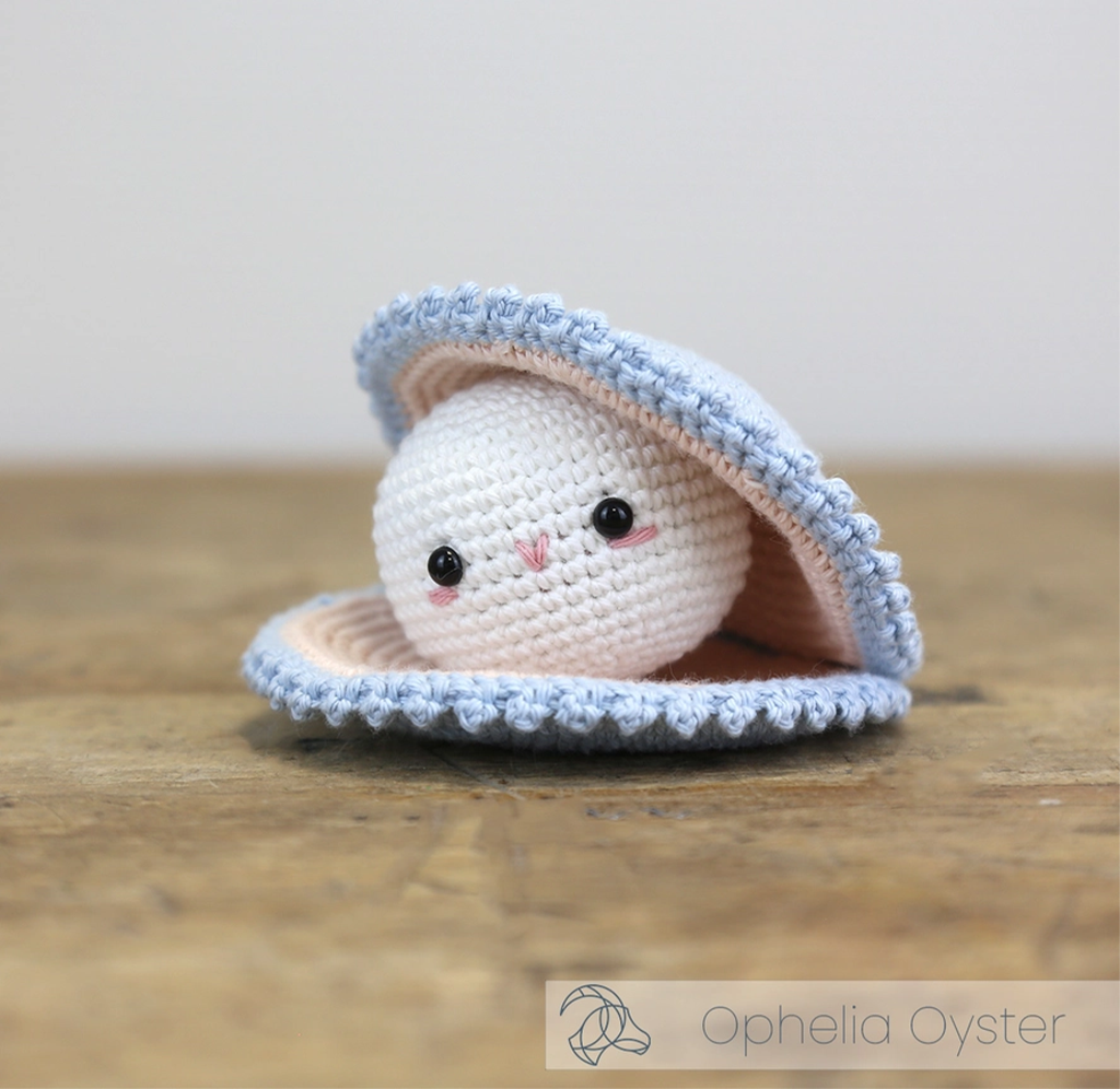 A cute crocheted white oyster character peeks from a pale blue, shell-shaped crocheted bed on a wooden table, with a shy, cozy expression.