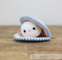 A cute crocheted white oyster character peeks from a pale blue, shell-shaped crocheted bed on a wooden table, with a shy, cozy expression.