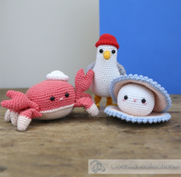 Crocheted coastal sea creatures—a red crab, white seabird in a red cap, and smiling shell—sit on a wooden surface against blue and gray walls, evoking a playful, handmade mood.