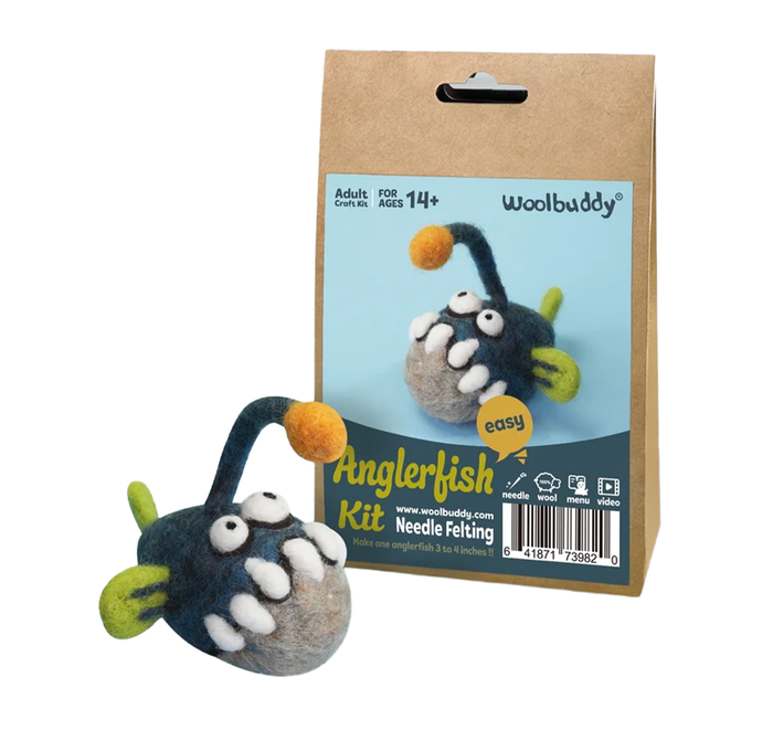 Woolbuddy Anglerfish needle-felting kit packaging beside a finished fuzzy fish with bulging eyes, green fins, and a dangling orange lure; playful craft display.