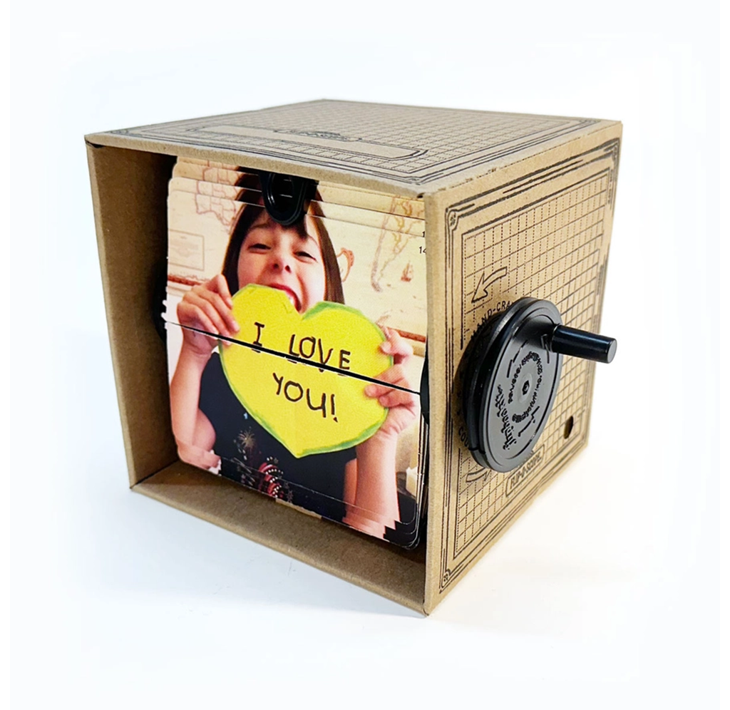 A handmade cardboard fliposcope displays a smiling child holding a yellow heart marked “I love you,” with a black side crank; playful, heartfelt tone.