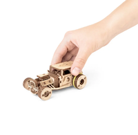 A hand gently races a small wooden vintage-style car on a white background, highlighting its intricate cutout details and handcrafted wheels.