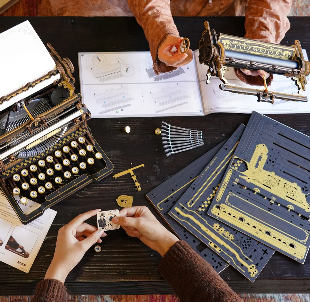 Overhead view of hands assembling a vintage-style typewriter from gold and navy parts, with an instruction booklet and finished typewriter nearby.