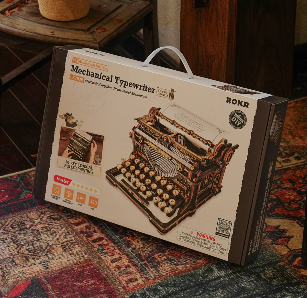 A ROKR mechanical typewriter model kit box rests on a patterned rug, showing an ornate vintage typewriter illustration and DIY kit details in a cozy room.