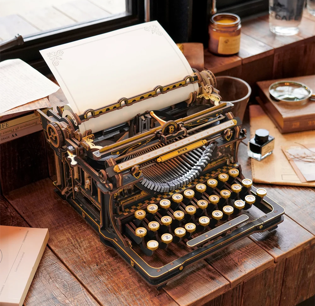 An ornate black-and-gold antique typewriter with a blank sheet of paper sits on a wooden desk by a window, evoking a warm, nostalgic writing mood.