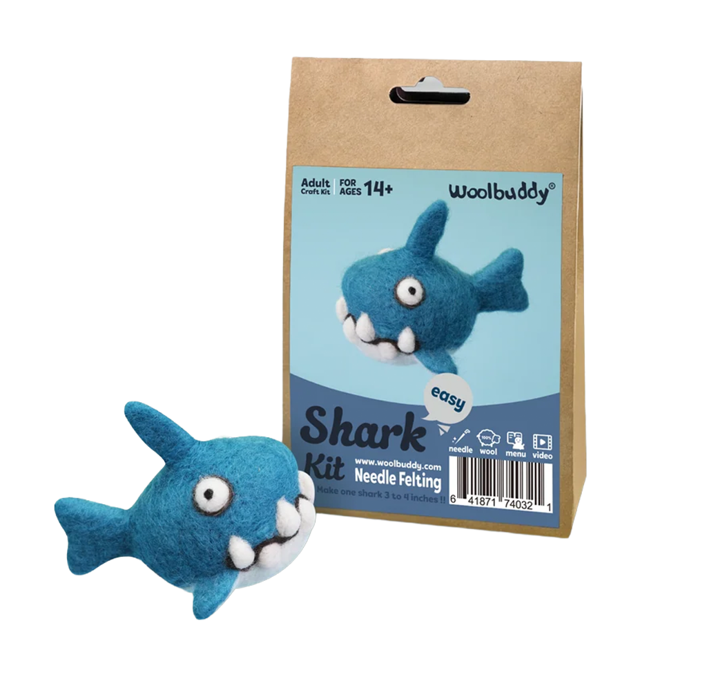 A cheerful blue felt shark sits beside a Woolbuddy needle-felting kit package featuring the same toothy character, on a clean white background.