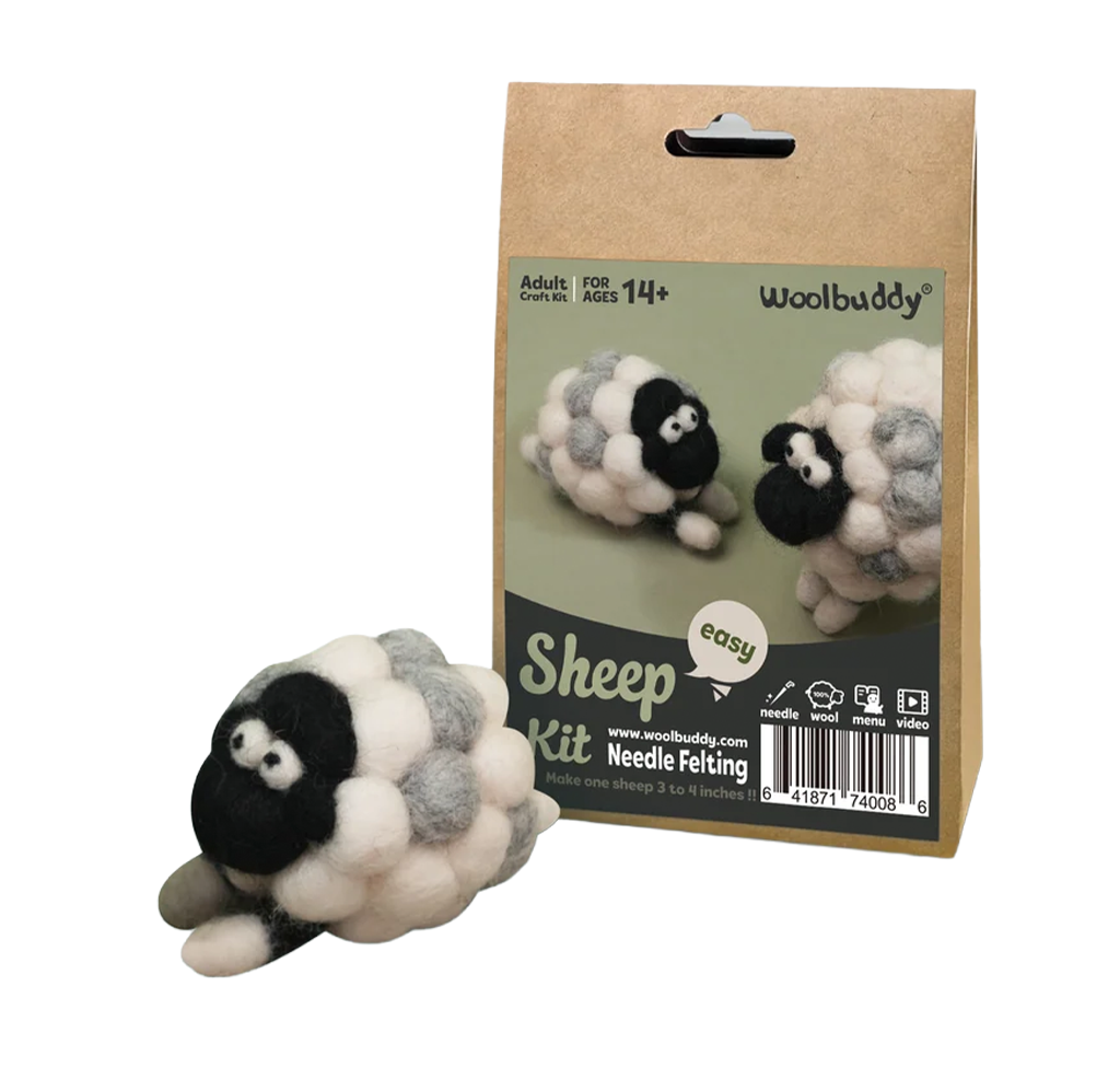 A Woolbuddy needle-felting sheep kit box stands behind a finished fluffy black, white, and gray sheep; the handmade craft display feels playful and inviting.